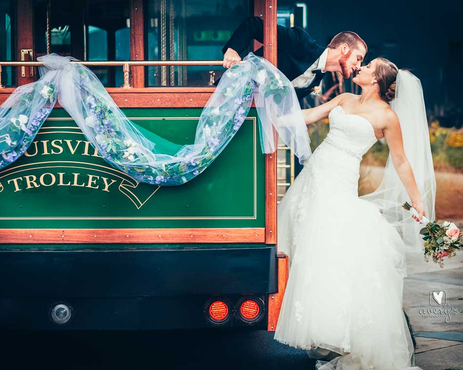 Bride and groom with a private trolley charter, leaning away from the green Toonerville trolley with a romantic kiss